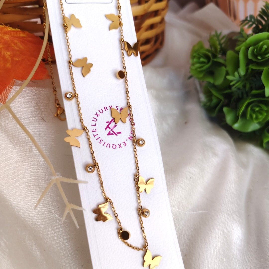 Gold butterfly necklace on a white card with a brand logo, placed on a textured surface with greenery in the background.